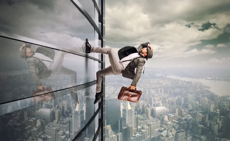 Businessman with hat and briefcase climbing the glass face of a skyscraper high above a city
