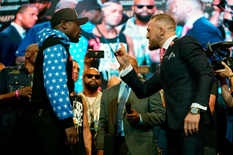 Conor McGregor in a pinstripe suit pointing at Floyd Mayweather, in a stars hoodie, at their press conference face-off