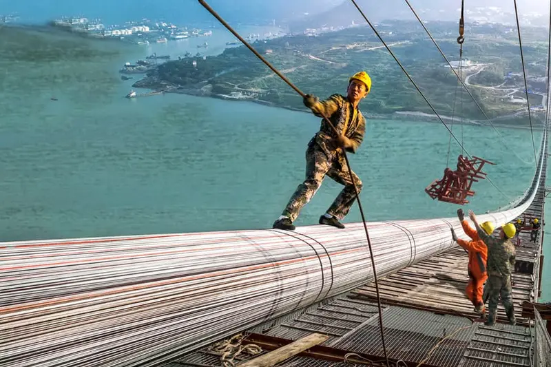 Construction workers in hard hats on the main cable of a suspension bridge high above a river