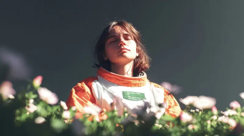 Young woman in an astronaut suit among flowers, eyes closed, face turned to the sun