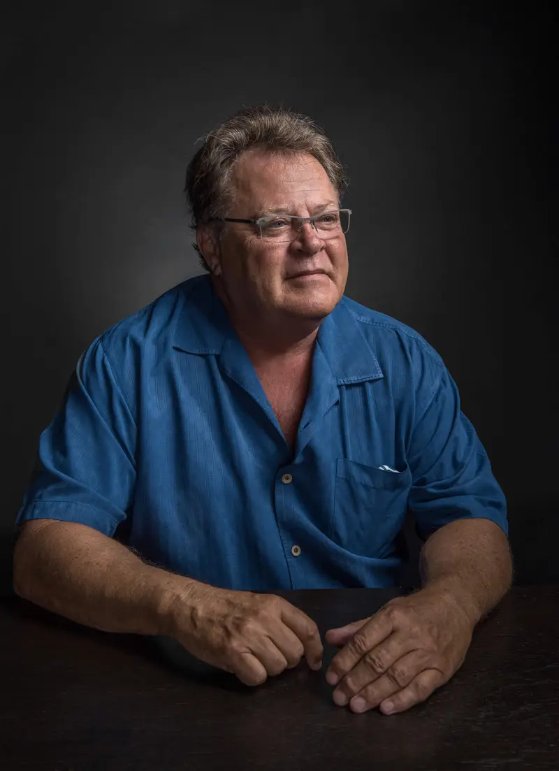 Portrait of an older man in a blue shirt, seated at a table and looking off to the side