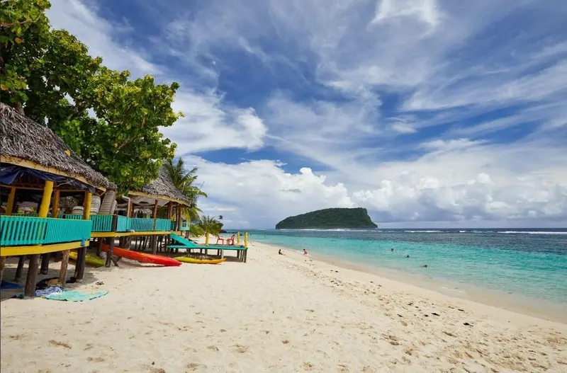 A quiet Samoan beach with thatched fales, kayaks on white sand and a small island offshore