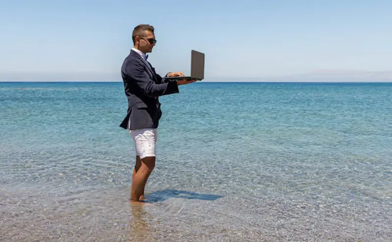 A businessman in a blazer and shorts stands ankle-deep in the sea, working on a laptop