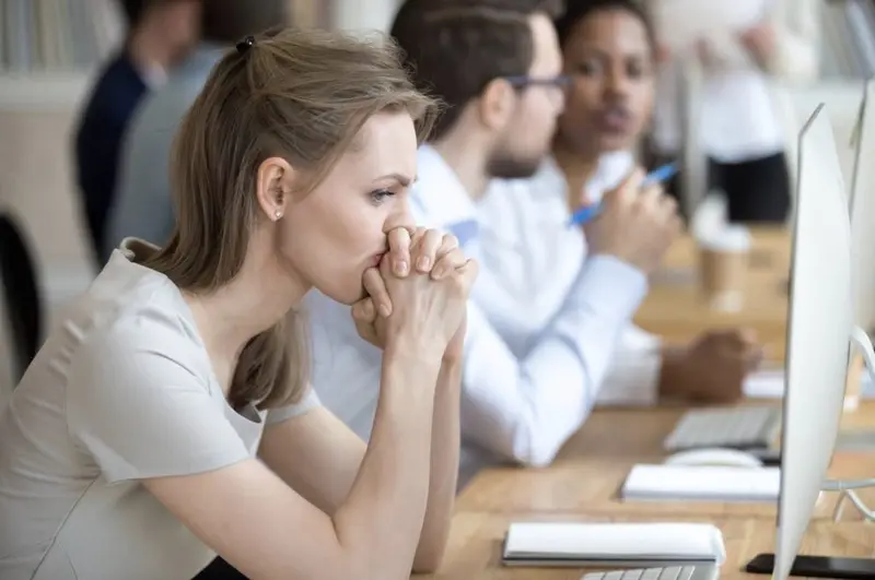 Anxious woman with hands clasped at her mouth at an office desk while colleagues talk beside her
